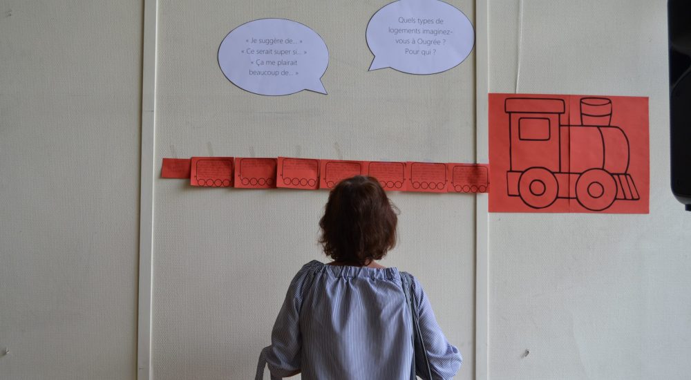 Personne face à un mur qui lis des phylactères où il est écrit : "Quels types de logements imaginez-vous à Ougrée ? Pour qui ?".