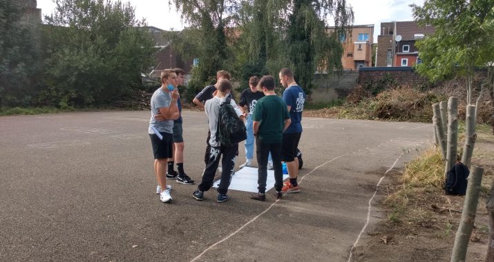 Groupe d'enfant au milieu d'une cour et en cercle autour d'une grande feuille blanche.