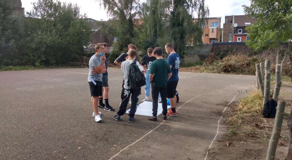 Groupe d'enfant au milieu d'une cour et en cercle autour d'une grande feuille blanche.