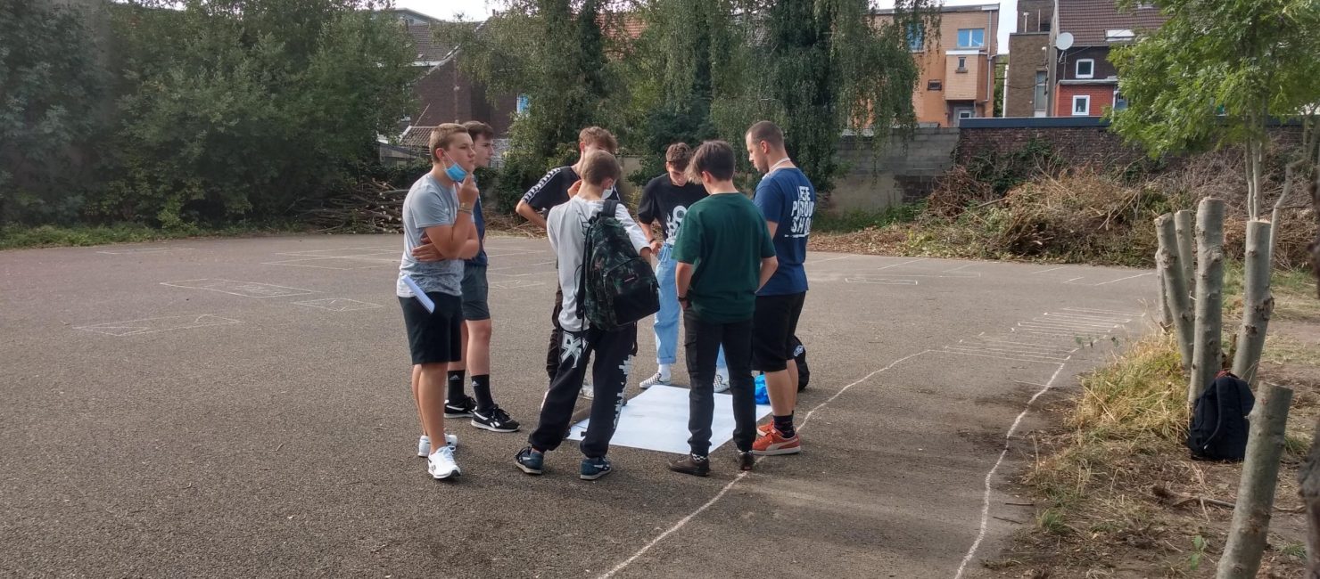 Groupe d'enfant au milieu d'une cour et en cercle autour d'une grande feuille blanche.